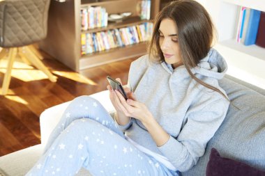 Yüksek açı çekim hoodie ve metin mesajlaşma koltukta evde dinlenirken giyen güzel genç kadın. 