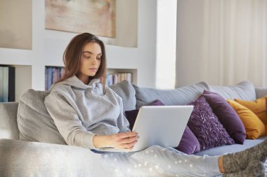 Atış süre kanepede rahatlatıcı ve onu laptop kullanarak hoodie ve pijama pantolon giyiyor rahat bir genç kadının.