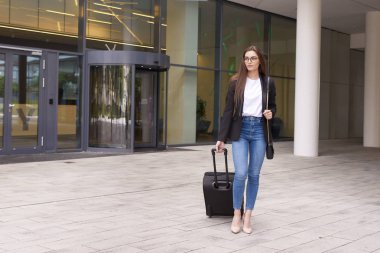 Bavuluyla modern ofis binasının yanında yürüyen çekici genç bir kadının tam boy fotoğrafı..