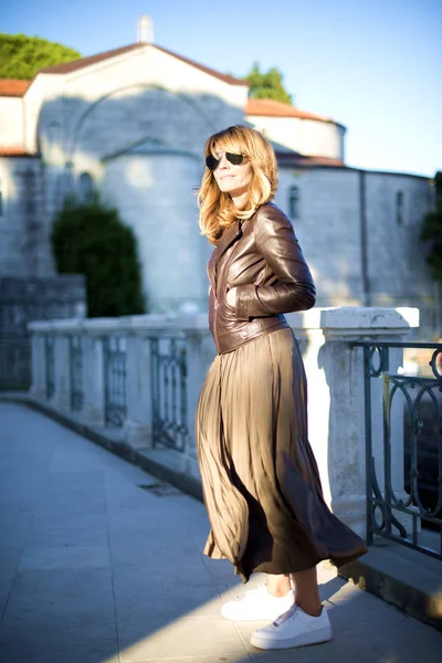 Full length shot of happy adult woman smiling outdoors and walking on city street. Pretty woman wearing sunglasses and biker jacket while relaxing outdoor.