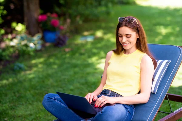 在花园里日光浴床上坐在土耳其座位上用笔记本电脑射击的年轻女子 — 图库照片