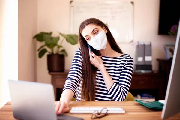 Shot of young businesswoman wearing casual clothes and face mask while working from home. Attractive female sitting at desk and making a call while working on laptop and computer. Home office.