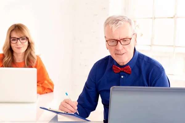 オフィスでラップトップを使用している間 蝶ネクタイと眼鏡をかけている上級ビジネスマンのショット 彼の後ろに座ってノートに取り組む中年の実業家 — ストック写真