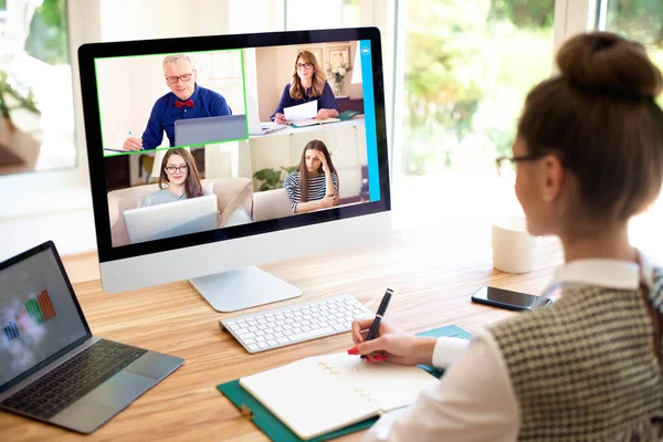 Back view of woman sitting behind her computer and having discussion and online meeting in video call with her collagues. Businesswoman working from home. Home office.