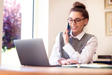 Bilgisayarının arkasında oturan mutlu genç kadının yüksek açılı görüntüsü ve evde çalışırken telefon görüşmesi yapması. Merkez Ofis. 