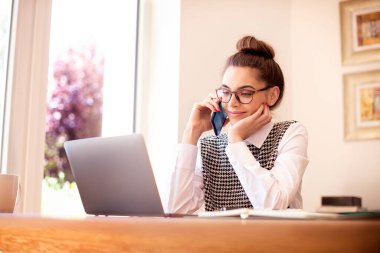 Bilgisayarının arkasında oturan mutlu genç kadının yüksek açılı görüntüsü ve evde çalışırken telefon görüşmesi yapması. Merkez Ofis. 