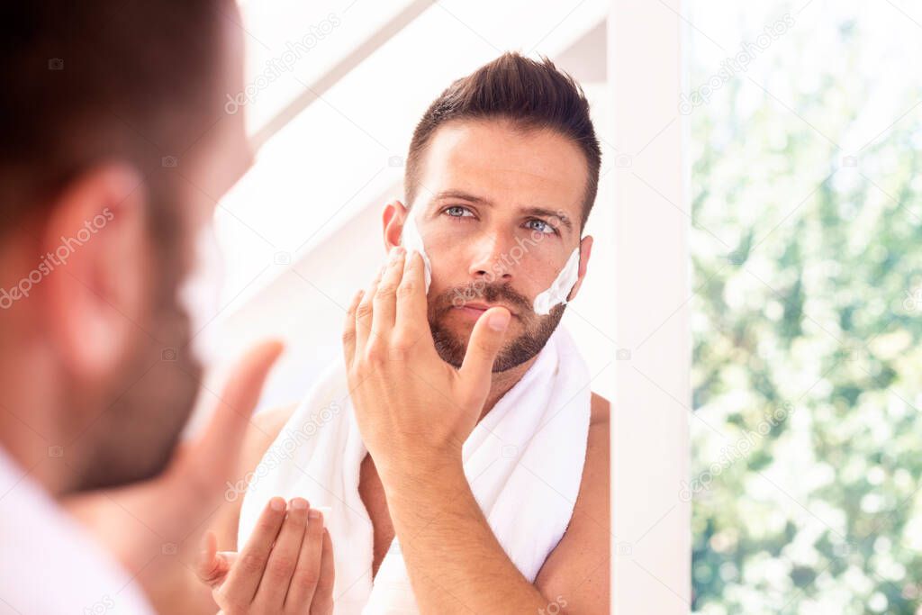 Close-up shot of handsome young man with towel in his neck standing in front of mirror and applying shaving foam to his face.