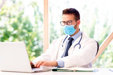 Yüz maskesi takan ve dizüstü bilgisayarda çalışan erkek doktorun portresi..