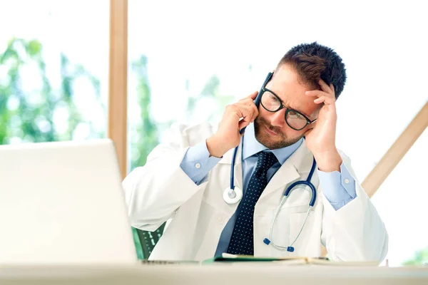 Shot of stressed male doctor sitting at desk behind his laptop and making a call
