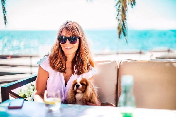 Orta yaşlı bir kadının güneşli bir günün tadını çıkarırken kral Charles Cavalier köpeğiyle deniz kenarında dinlenirken çekilmesi.. 