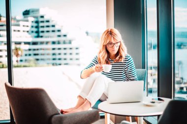 Evde dizüstü bilgisayarla çalışırken kendine güvenen bir kadının sabah kahvesini içmesi. Merkez Ofis. 