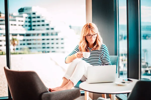 Evde dizüstü bilgisayarla çalışırken kendine güvenen bir kadının sabah kahvesini içmesi. Merkez Ofis. 