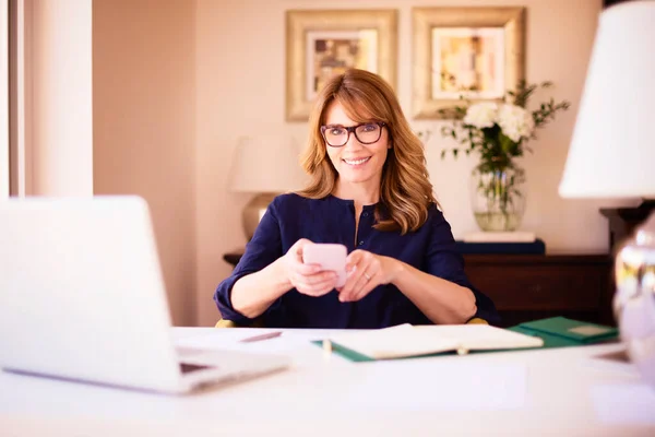 在家里工作的时候 坐在笔记本电脑后面的一位成熟的女商人坐在办公桌前 一边发短信一边笑 总部办公室 — 图库照片