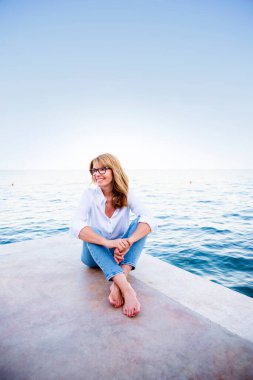 Sahilde oturup rahatlarken beyaz gömlek ve kot pantolon giyen mutlu bir kadının yüksek açılı fotoğrafı..