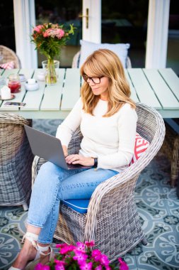 Evde balkonda otururken bilgisayarını kullanarak gülümseyen olgun bir kadının yüksek açılı görüntüsü. Merkez Ofis. 