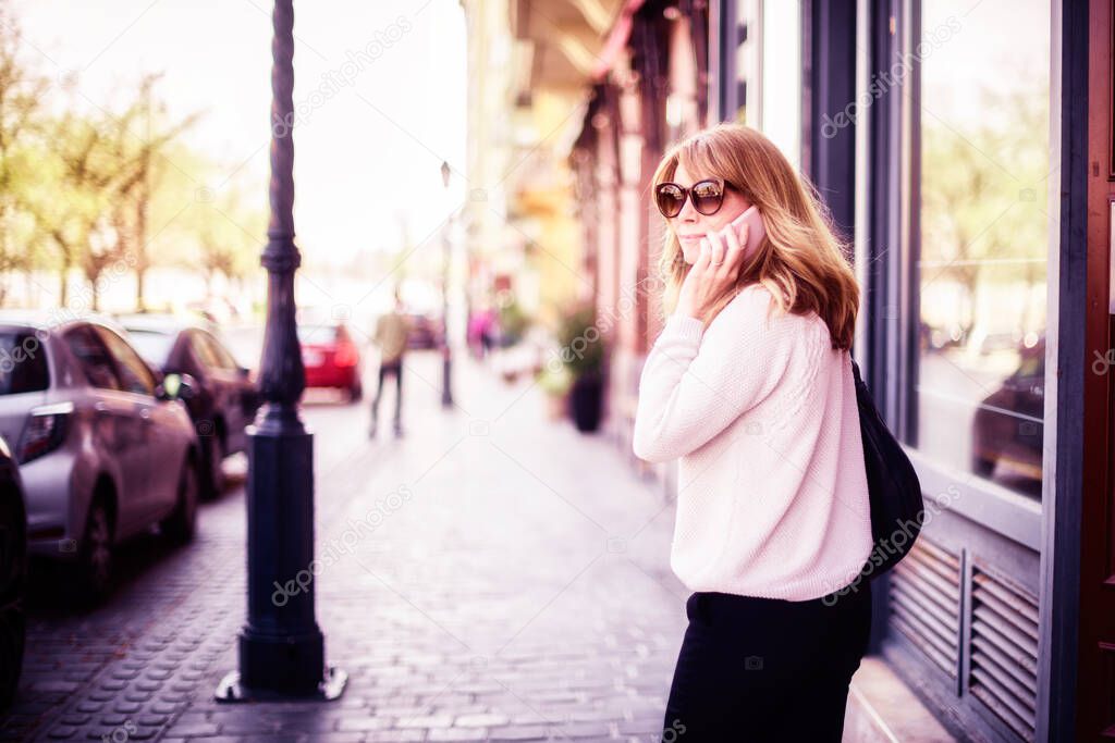 Shot of middle aged woman wearing casual clothes and sunglasses and using mobile phone while walking on the street.