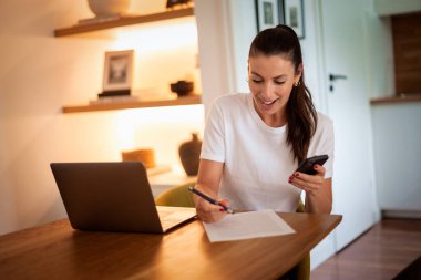 Çekici esmer saçlı kadın modern evinde oturuyor ve çalışıyor. Akıllı telefon ve dizüstü bilgisayar kullanan orta yaşlı bir kadın. Sıradan kıyafetler giyen çekici bir iş kadını. Merkez Ofis. 