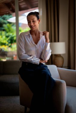 Güzel, orta yaşlı bir kadının koltukta oturup hayal kurduğu bir fotoğraf. Kumral kadın, beyaz gömlek ve kot pantolon giyiyor..