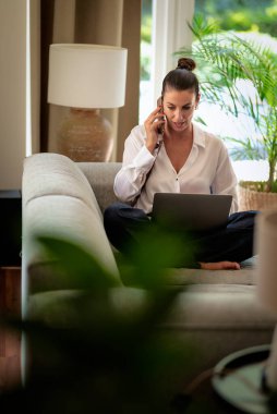 Kendine güveni tam orta yaşlı bir kadın evde uzaktan çalışıyor telefonda konuşurken gülümsüyor ve dizüstü bilgisayar kullanıyor. Sıradan görünüş ve modern iç mekan. Merkez Ofis.