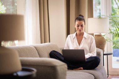 Kendinden emin orta yaşlı bir kadın evden uzakta çalışıyor. Dizüstü bilgisayar kullanırken gülümsüyor. Gündelik görünüş, rahat ve modern iç mekan. Merkez ofis. Serbest çalışma..