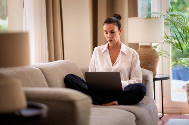 Kendinden emin orta yaşlı bir kadın evden uzakta çalışıyor. Bir dizüstü bilgisayar kullanırken düşünceli görünüyorsun. Gündelik görünüş, rahat ve modern iç mekan. Merkez ofis. Serbest çalışma..