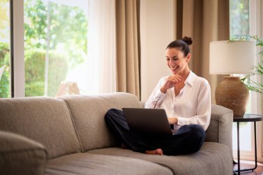 Kendinden emin orta yaşlı bir kadın evden uzakta çalışıyor. Dizüstü bilgisayar kullanırken gülümsüyor. Gündelik görünüş, rahat ve modern iç mekan. Merkez ofis. Serbest çalışma..