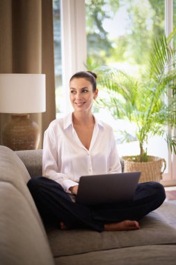 Kendinden emin orta yaşlı bir kadın evden uzakta çalışıyor. Dizüstü bilgisayar kullanırken gülümsüyor. Gündelik görünüş, rahat ve modern iç mekan. Merkez ofis. Serbest çalışma..