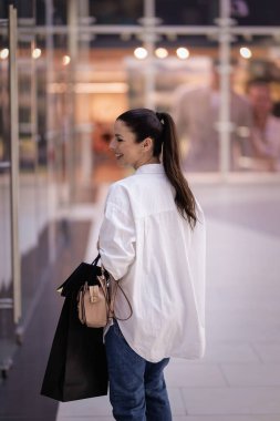 Alışveriş merkezinde yürüyen ve vitrinlere bakan orta yaşlı bir kadın. Kumral kadın, beyaz gömlek ve kot pantolon giyiyor.. 