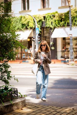 Gündelik giysiler içinde orta yaşlı stil sahibi bir kadın güneşli bir günde şehir caddesinde yürüyor. Elinde kağıt kahve fincanı tutan ve tekstil torbası taşıyan kendine güvenen bir kadın. Tam uzunluk. 