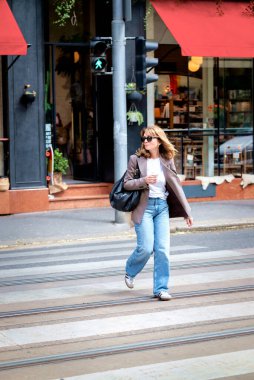 Sıradan giysiler içinde orta yaşlı stil sahibi bir kadın yaya geçidinden karşıya geçiyor. Kendine güvenen bir kadın elinde bir kahve fincanı tutuyor ve çanta taşıyor. Tam uzunluk. 