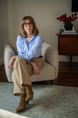 Koltukta oturan, omuz hizasında sarı saçlı ve şık gözlükleri olan kendinden emin orta yaşlı bir kadın. Mavi gömlekli çekici bir kadın ve pozitif bir gülümsemeyle kameraya bakıyor. Tam uzunluk.