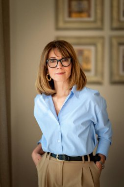 Kendine güvenen, orta yaşlı, omuz hizasında, sarı saçlı ve şık gözlükleri olan bir kadın. Mavi gömlekli çekici bir kadın ve pozitif bir gülümsemeyle kameraya bakıyor. Yakın plan..