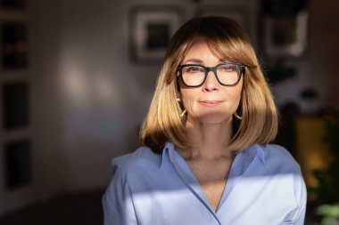 Kendine güvenen, orta yaşlı, omuz hizasında, sarı saçlı ve şık gözlükleri olan bir kadın. Mavi gömlekli çekici bir kadın ve pozitif bir gülümsemeyle kameraya bakıyor. Yumuşak ışıklandırma, bulanık arkaplan. Yakın plan..