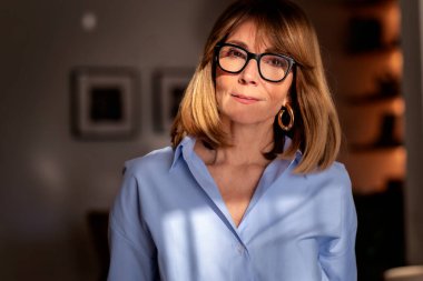 Kendine güvenen, orta yaşlı, omuz hizasında, sarı saçlı ve şık gözlükleri olan bir kadın. Mavi gömlekli çekici bir kadın ve pozitif bir gülümsemeyle kameraya bakıyor. Yumuşak ışıklandırma, bulanık arkaplan. Yakın plan..