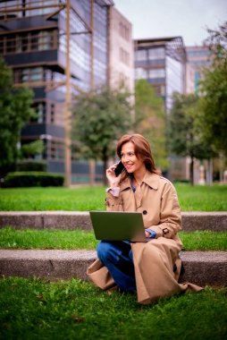 Sonbahar günü dizüstü bilgisayarıyla parkta oturan orta yaşlı bir kadın. Trençkot ve kot pantolon giyen kendinden emin bir kadın. Uzaktan kumanda. Tam uzunluk. 