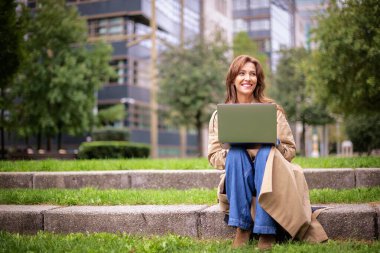 Sonbahar günü dizüstü bilgisayarıyla parkta oturan orta yaşlı bir kadın. Trençkot ve kot pantolon giyen kendinden emin bir kadın. Uzaktan kumanda. Tam uzunluk. 