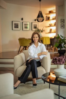 Güzel, gülümseyen orta yaşlı bir kadının koltukta oturup kameraya baktığı bir fotoğraf. Beyaz gömlek ve pantolon giyen esmer kadın. Tam boy çekim.