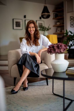 Güzel, gülümseyen orta yaşlı bir kadının koltukta oturup kameraya baktığı bir fotoğraf. Beyaz gömlek ve etek giyen esmer kadın. Tam boy çekim.