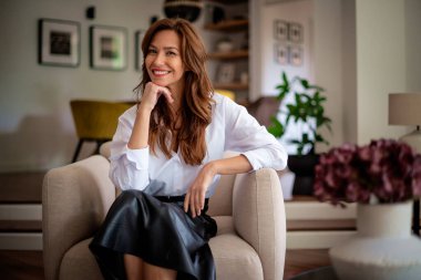 Güzel, gülümseyen orta yaşlı bir kadının koltukta oturup kameraya baktığı bir fotoğraf. Esmer kadın, beyaz gömlek ve etek giyiyor..