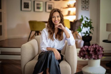 Güzel, gülümseyen orta yaşlı bir kadının koltukta oturup kameraya baktığı bir fotoğraf. Esmer kadın, beyaz gömlek ve etek giyiyor..