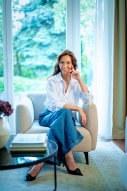 40 'larında şık esmer bir kadın, beyaz gömlek ve kot pantolon giyiyor ve pencerenin önünde oturuyor. Çekici bir kadın, esmer saçlı ve kendine güvenen bir gülümsemesi var. Tam boy çekim. 