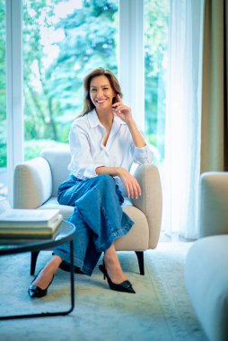 40 'larında şık esmer bir kadın, beyaz gömlek ve kot pantolon giyiyor ve pencerenin önünde oturuyor. Çekici bir kadın, esmer saçlı ve kendine güvenen bir gülümsemesi var. Tam boy çekim. 