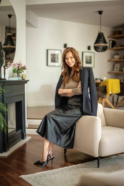 Güzel, gülümseyen orta yaşlı bir kadının koltukta oturup kameraya baktığı bir fotoğraf. Ceket ve etek giyen esmer saçlı kadın. Tam boy çekim.