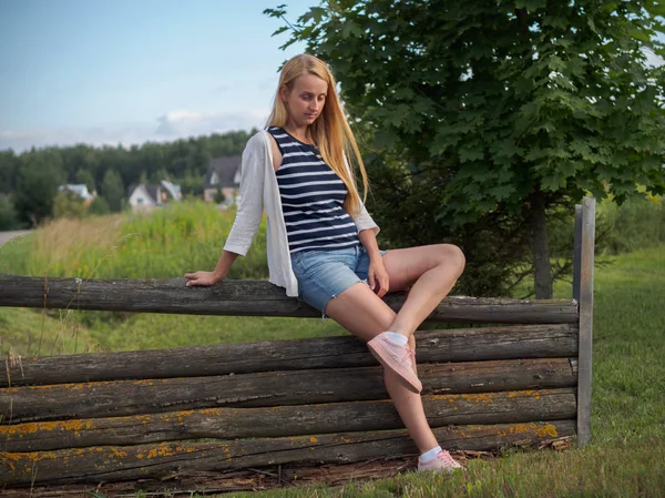 Cute blonde woman posing on old wooden fence in denim shorts — Stock Photo, Image