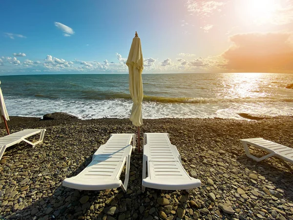 Plastic witte ligstoelen met een gesloten parasol op een leeg strand. — Stockfoto
