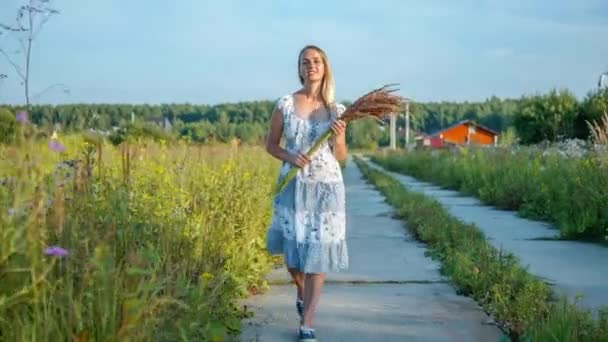 Blondynka w długiej sukience z kwiatami spacerująca po wiejskim krajobrazie — Wideo stockowe