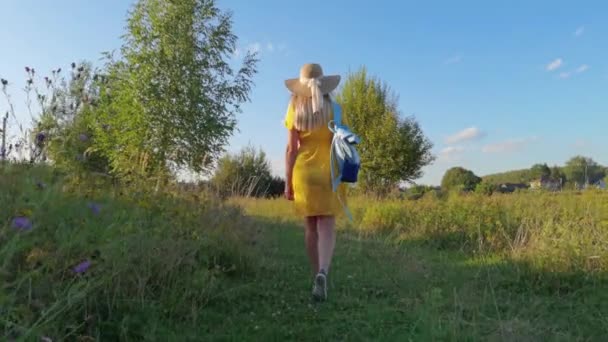 Женщина-путешественница в жёлтом платье с рюкзаком, идущая по лугу — стоковое видео