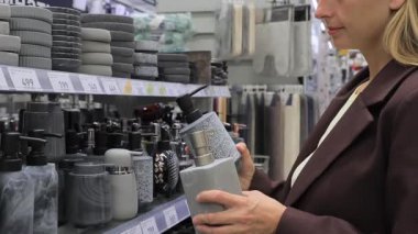 Kahverengi ceketli zarif bir kadın ev dekorasyon mağazasının banyo aksesuarları bölümünden şık gri bir seramik sabun makinesi seçiyor. Modern banyo tasarımı, iç dekorasyon, fonksiyonel stil ve ev gereçleri alışverişi kavramı.