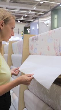 Sarı gömlekli genç bir kadın hırdavatçıda renkli sıcak hava balonu desenli duvar kağıdı rulosu seçiyor. İç dekorasyon, şık duvar kaplaması, yenileme projesi, modern tasarım çözümü ve ev dekorasyonu alışverişi kavramı.