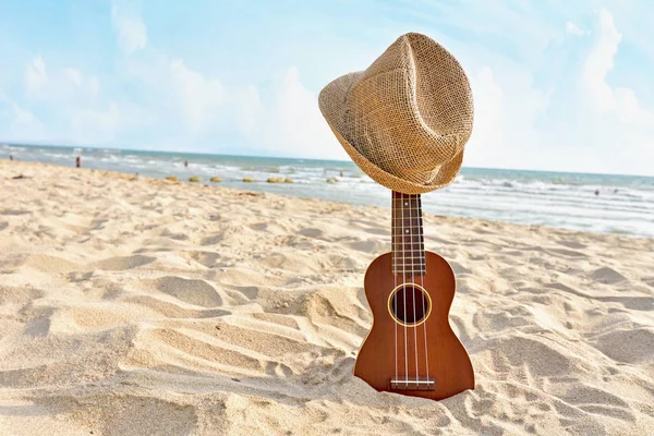 Акустическая гитара укулеле, стоящая на песчаном пляже в шляпе — стоковое фото